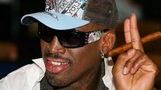 Dennis Rodman smiles and smokes a cigar wearing a baseball cap.