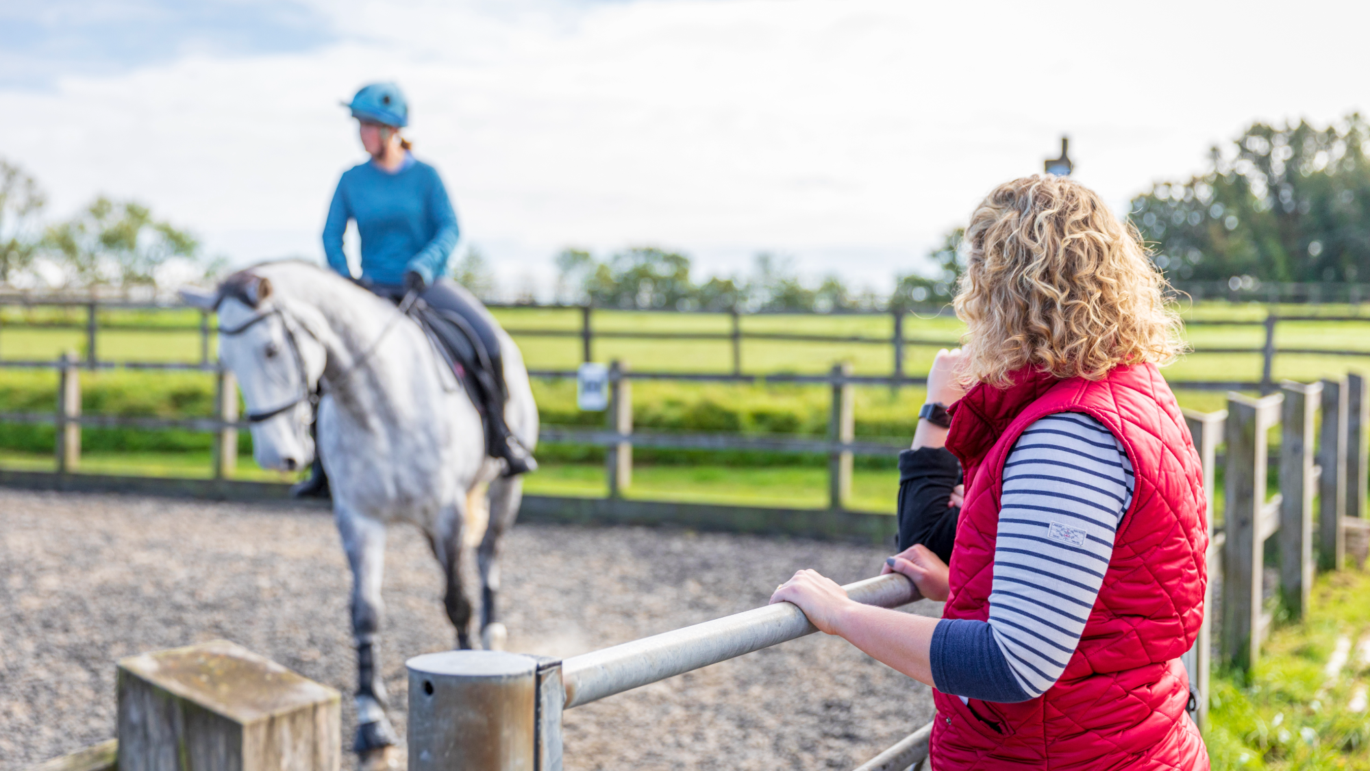 Two women watch a womean wearing blue riding a grey horse in an outdoor arena