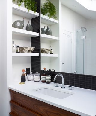 Bathroom basin and tap with black tiled splashback, white work surface, wood cabinetry, built in shelving and large mirror