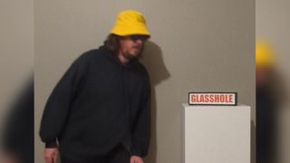 A person in a yellow bucket hat wearing Meta Ray-Ban smart glasses, approaching a sign that says "glasshole"