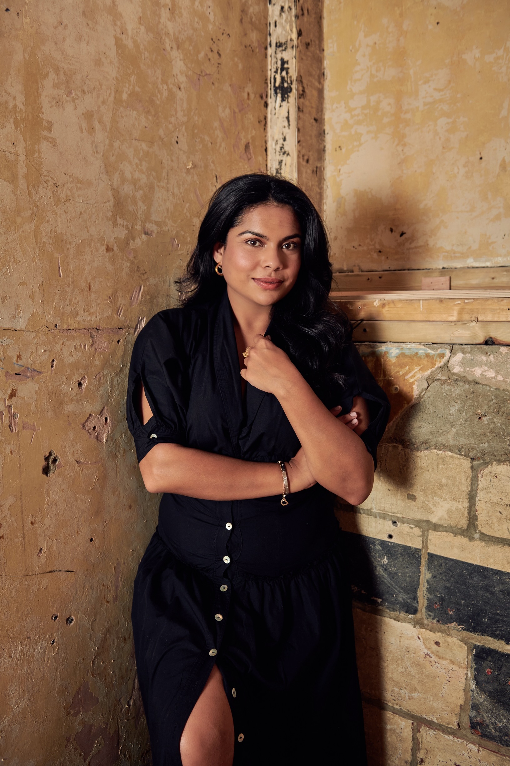 A woman in a buttoned black maxi dress with short sleeves poses with her dark hair left hanging on her shoulders against a bare cement and brick ochre wall.