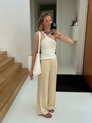 Anouk Yve taking a mirror selfie wearing wide-leg pants, a one-shoulder tank, beaded necklace, flip-flops, and carrying an oversize creamy leather tote.