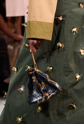 A snakeskin purse is shown in a close-up photo of a model walking down Ulla Johnson's spring 2025 runway show, carrying a python clutch bag with an olive green embellished trench