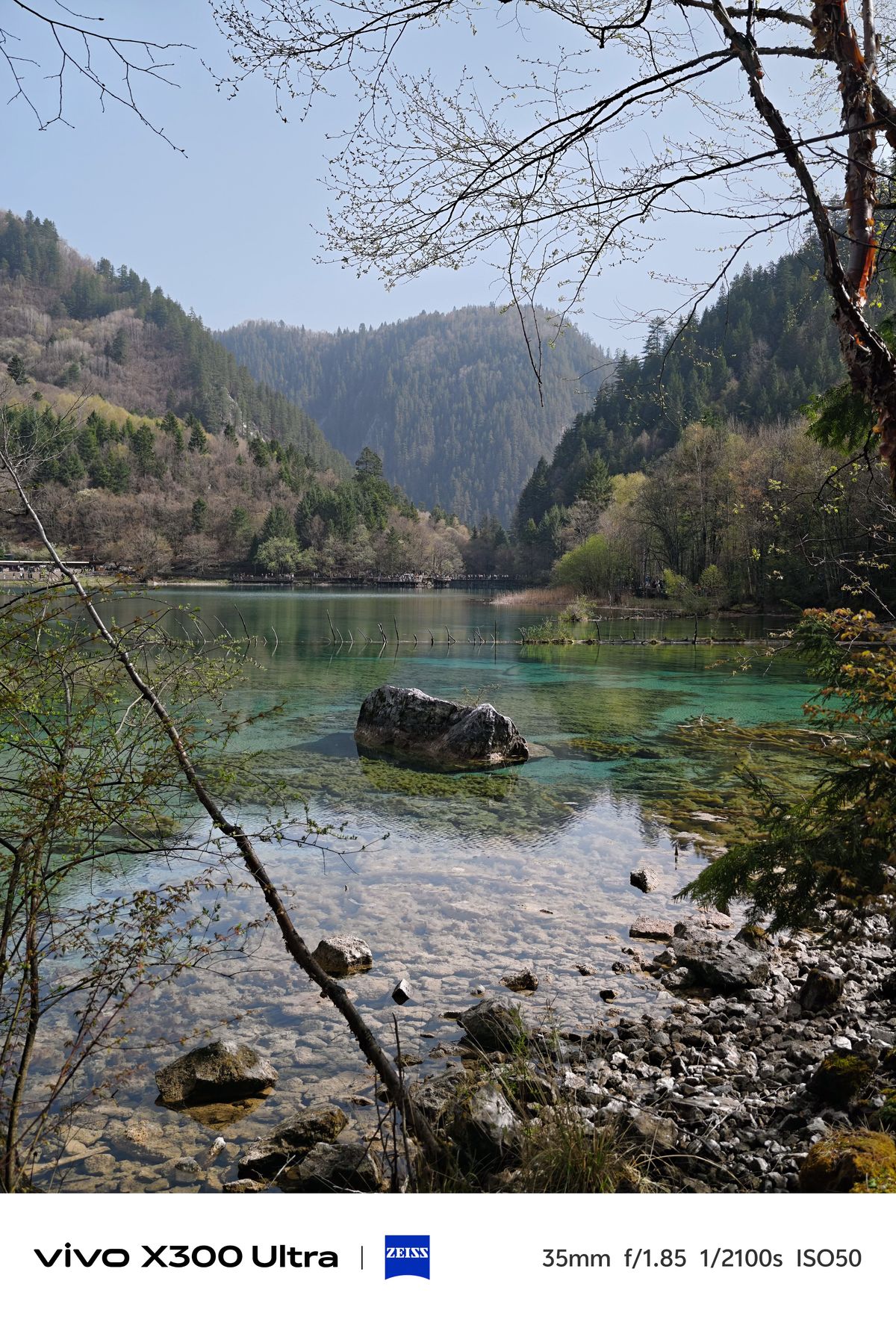 A clear lake shoreline with smooth stones below the surface, a central boulder, and forested mountains.