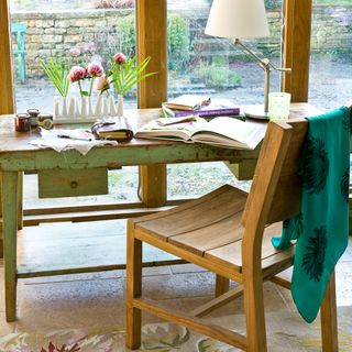 a distressed green wooden desk with books, a lampt and flowers on top, with a wooden chair, in a conservatory that overlooks a garden patio