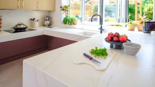 pale corian worktops with red kitchen cabinets below and wooden cabinets above as well as large bifold kitchen window above kitchen sink