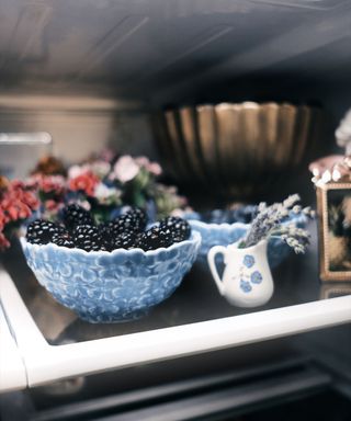 A fridge with a blue bowl of blackberries and a small white vase with flowers in it