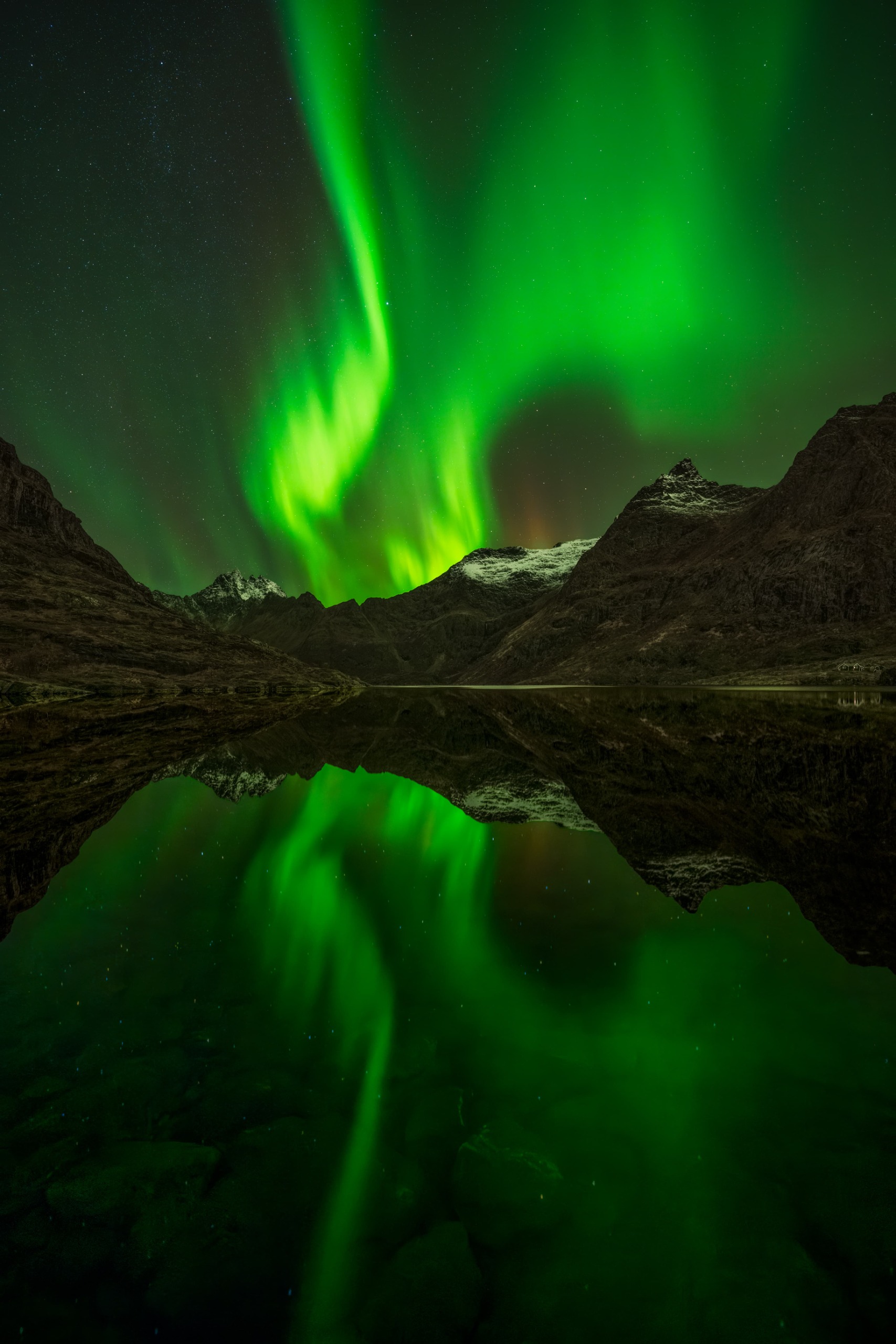 The Aurora Borealis dances above the mountains of &amp;Aring; in Norway's Lofoten Islands, its vibrant green light reflected across the calm waters below.