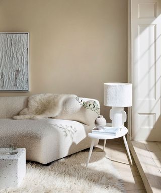Calming, cream and white living space, cream painted walls, curved boucle sofa with sheepskin throw, white wooden side table with textured, geometric table lamp, light wooden flooring, shaggy sheepskin cream rug