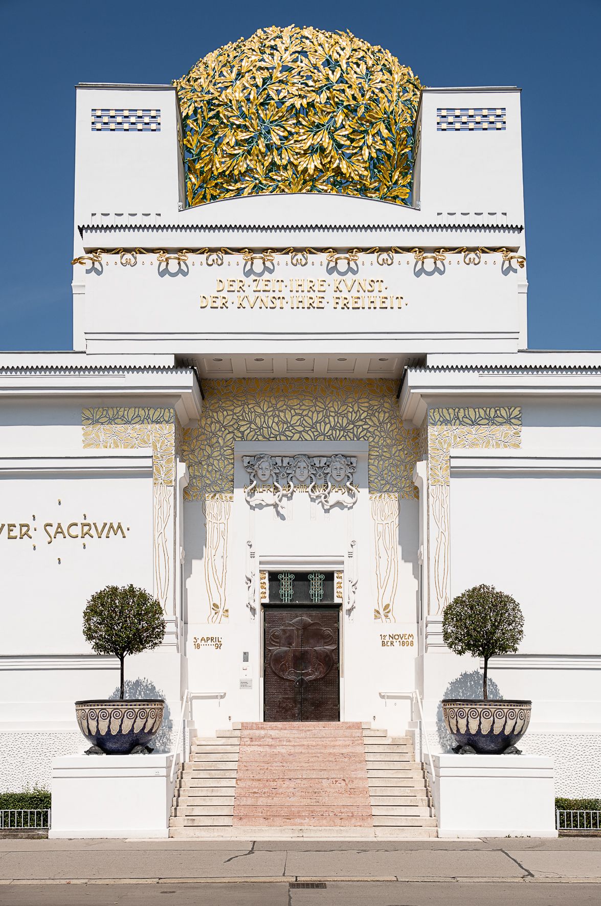 An historical building&#039;s golden-leaf frieze depicts a human procession characterized by a fantastical atmosphere and repeated floral patterns, while the rest of the facade is white.