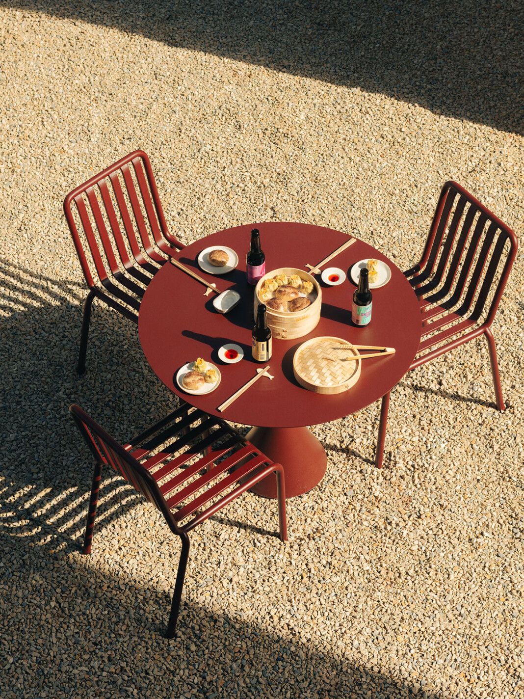 The sun-lit exterior of a French hotel boasts color-blocked outdoor iron furniture painted in a dark terracotta shade, topped with an Asian takeaway.