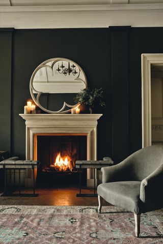 black living room with large round mirror, open fire, vintage rug, grey armchair, lit candles on mantel and vase of foliage