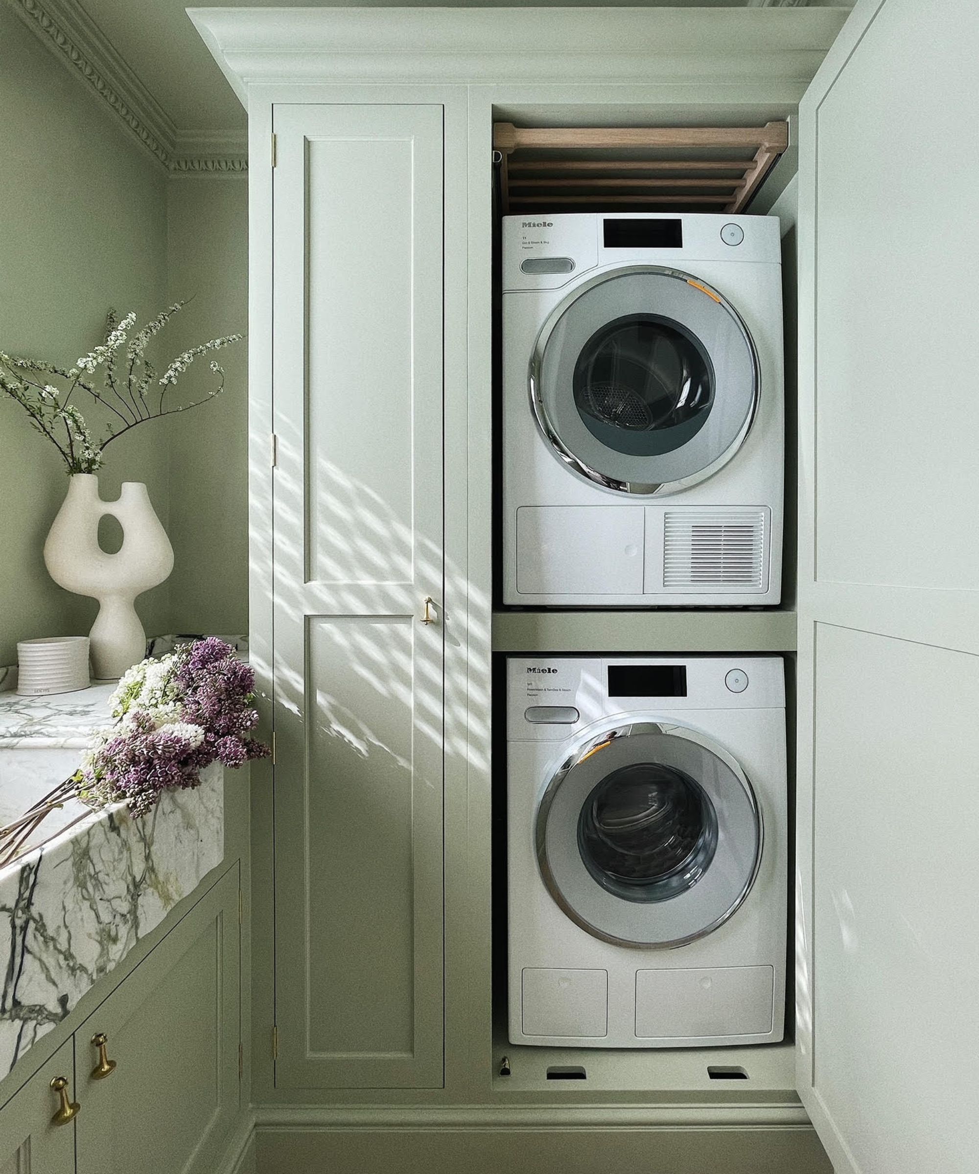 Stacked washing machine and dryer built into sage green cabinetry, with a marble sink and fresh flowers on the counter in a softly lit utility room.