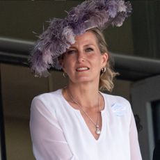 Duchess Sophie wears a sheer white shirt and large purple hat featuring lots of feathers, and husband Prince Edward stands beside her