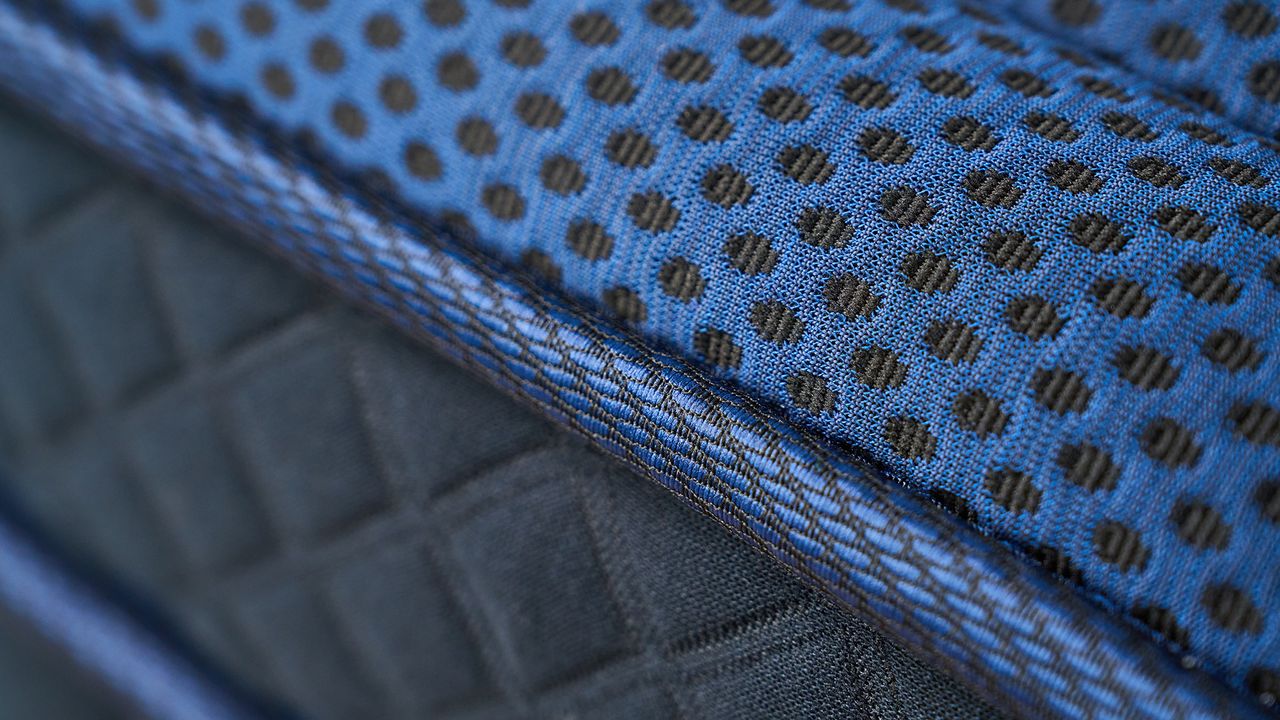 The image shows the close-up details of the DreamCloud Ultra Hybrid, a navy blue mattress placed on a white bed frame in a neautrally decorated bedroom