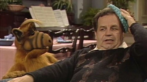 ALF the furry, croissant alien is looking at a man who is slumped on the couch holding an ice pack to their head.