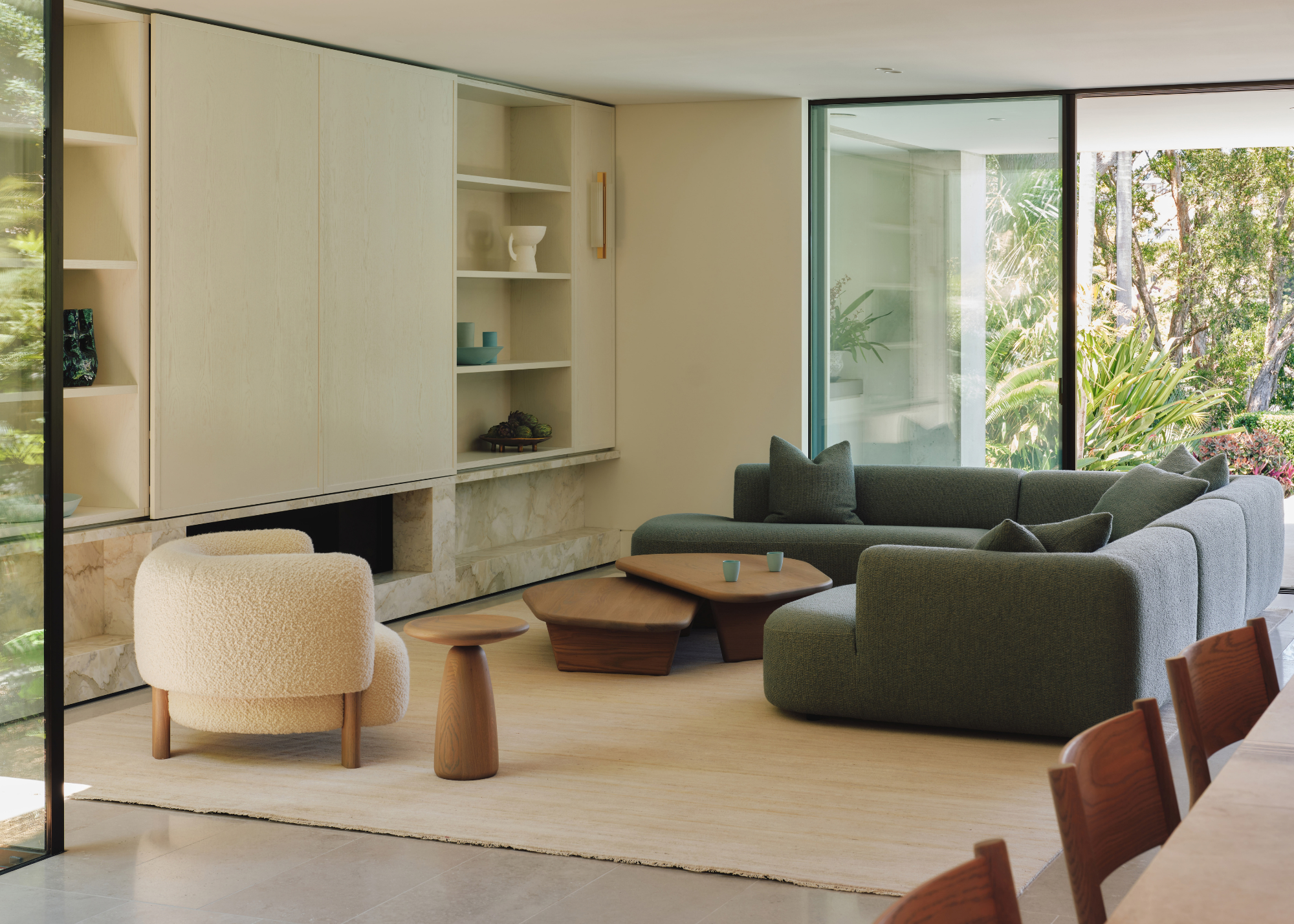 A bright living room with a green couch, a set of coffee tables, a stool, and a white bucle armchair, beside a TV unit