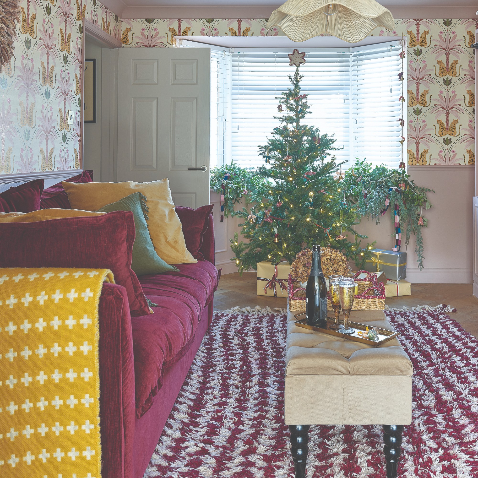 A living room with a floral wallpaper and a burgundy sofa and a decorated Christmas tree with presents by the window