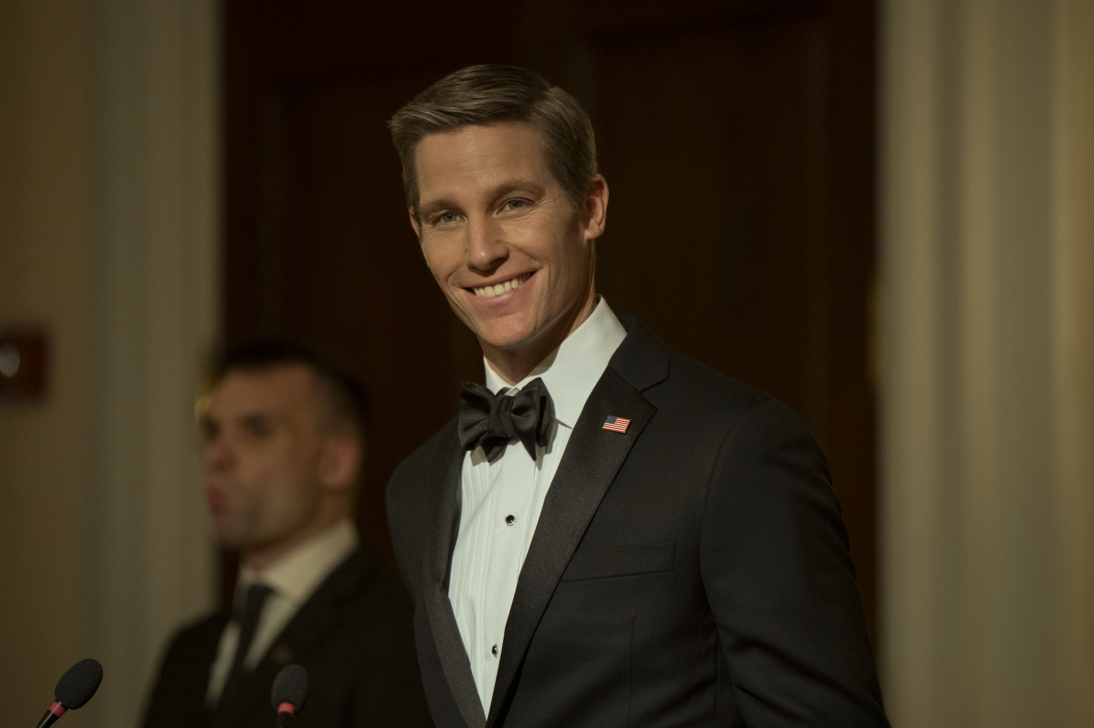 Ward Horton as Governor Richard Hagan wearing a suit and bow tie smiling while giving a speech in the night agent season 3