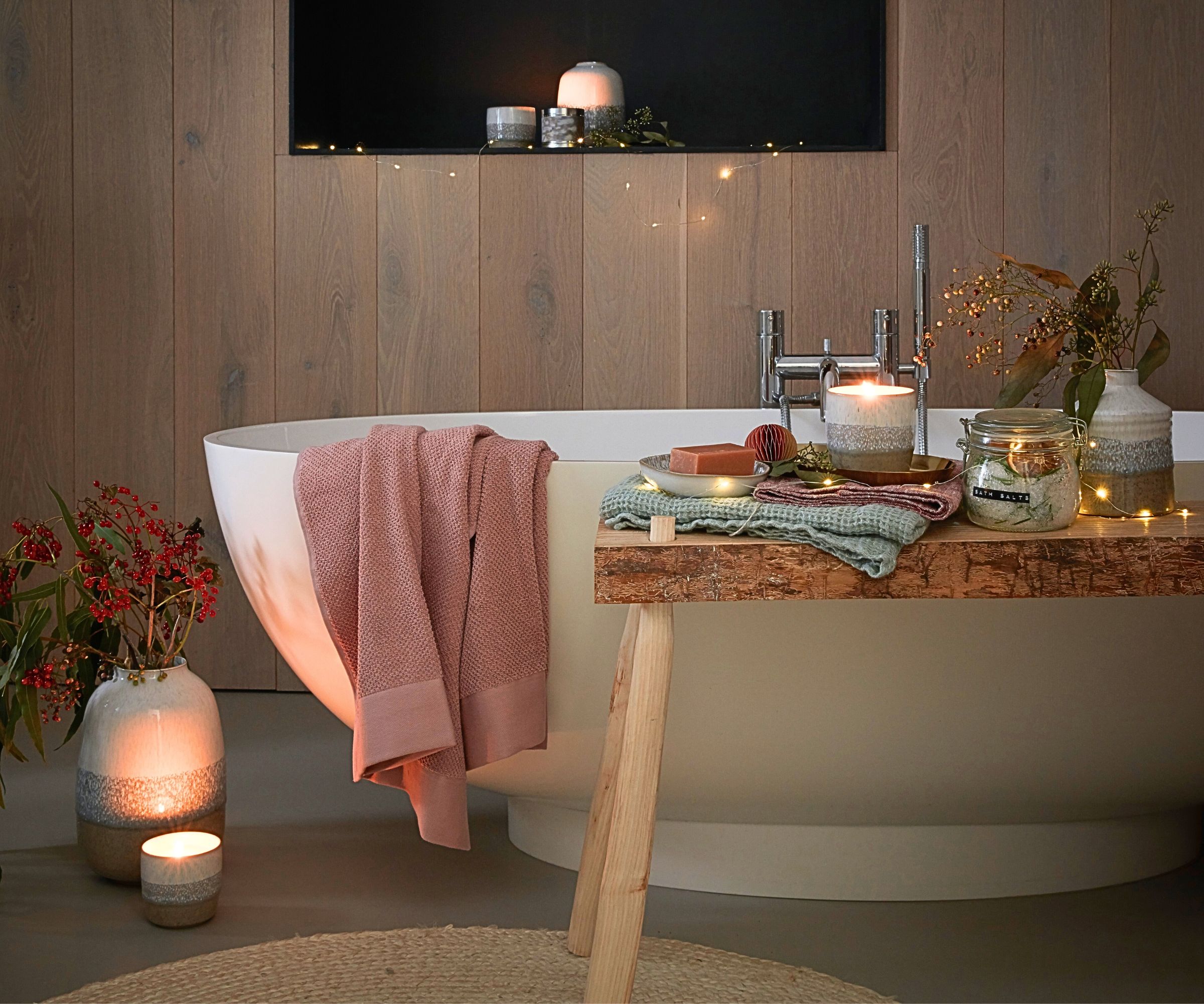A cosy, candlelit bathroom styled for winter with a white freestanding oval bath against oak wall panelling. A rustic wooden bath rack holds folded towels, bath salts and candles, while fairy lights, red berry stems and ceramic vases create a warm, festive atmosphere.