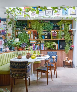 Characterful basement kitchen with skylight, ceiling and walls decorated with plants, photos, artwork and books, wooden shelving unit, green bench seating, rounded marble tulip dining table, wooden seats, wooden flooring