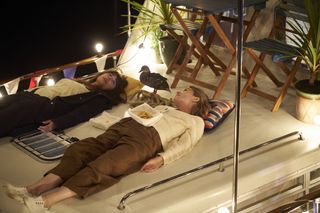 Esther (Zahra Ahmadi) and Martha (Sally Bretton) are lying on their backs on the roof of the houseboat. It is night, but the strings of lights all around them are switched on. Selwyn the duck is standing between them on a small pile of rope. There is an empty box of chips lying in between them, and a half-eaten box of chips sitting on Martha's stomach