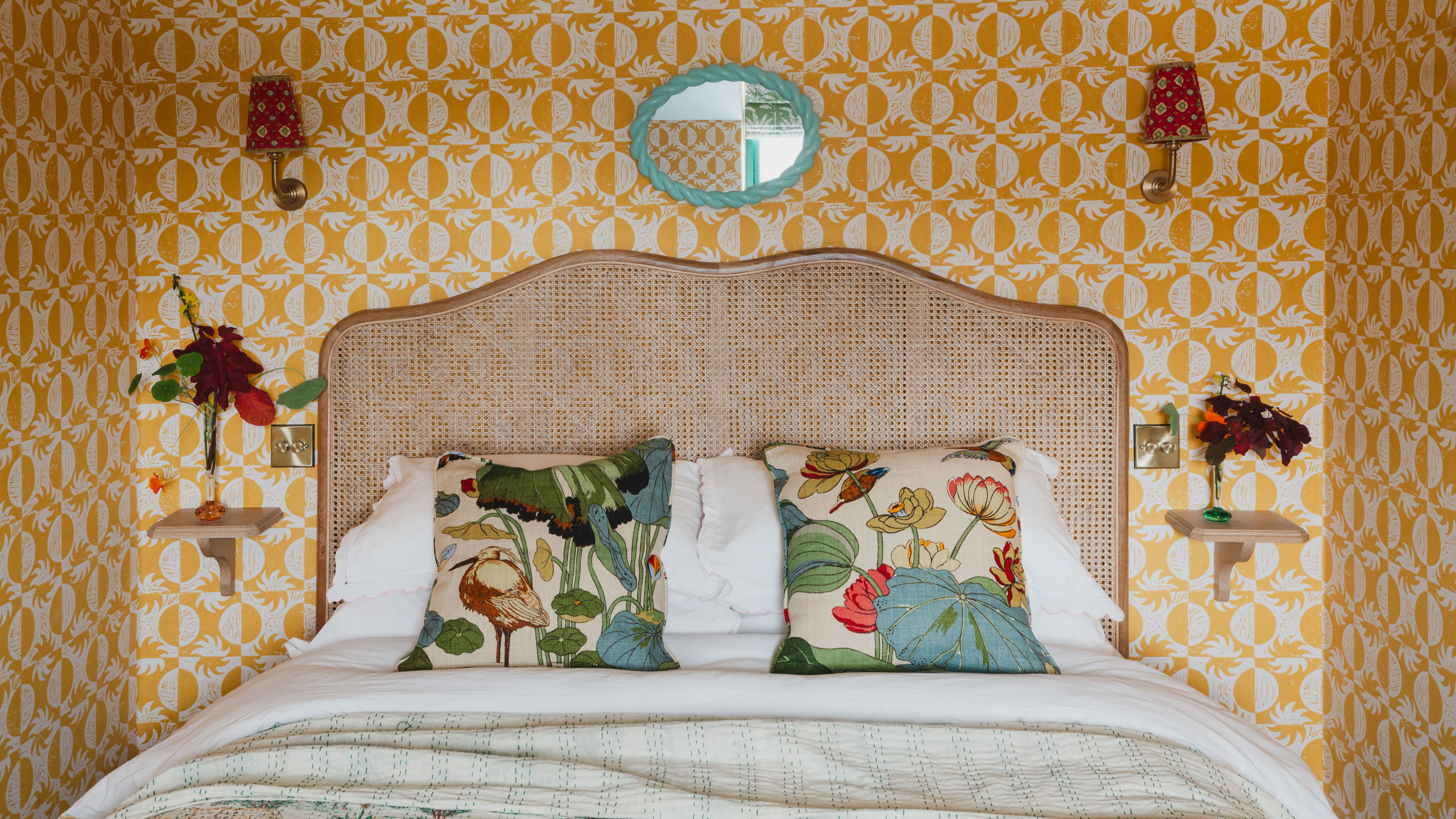 Yellow bedroom wallpaper with rattan headboard, turquoise mirror and floral throw cushions
