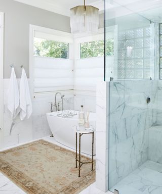 White and gray bathroom space with marble tiles on the floor and on the walls, neutral, textured rug, shower with glass door, glass and gold hanging pendant, towels on hooks on wall, windows with white fitted blinds, metal slender side table positioned next to white bath