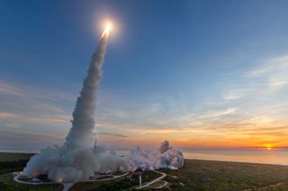 A conical column of smoke trails upward from the ground, where it shoot wide, toward a blindingly bright stretch of fire in the sky, connected to the end of a faint, small rocket. the grassy area surrounding the launchpad gives way to the seashore in the background as the orange sun peaks through a strip of cloud on the horizon.