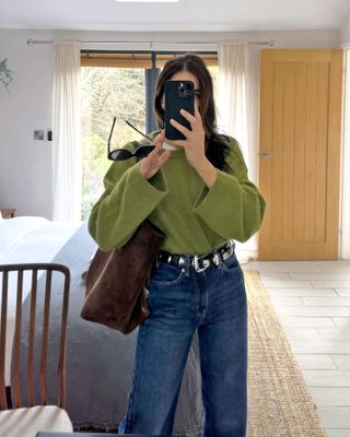 Die britische Mode-Influencerin Jessica Skye posiert für ein Spiegel-Selfie in einem grünen Pullover mit Rundhalsausschnitt, einer braunen Wildleder-Einkaufstasche, einem mit Nieten besetzten Khaite-Gürtel und Jeans