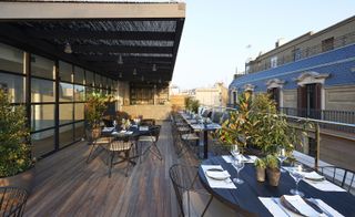 View of a rooftop dining area at The Serras featuring wooden decking, round dark wood tables with tableware, black chairs, green plants, a bar and a view of nearby buildings