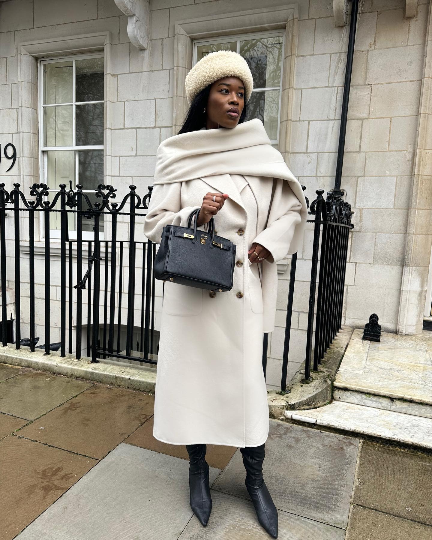 @nlmarilyn wearing a cream coat with an attached scarf, black bag, shearling hat, and boots.