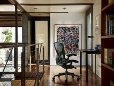 A deep green Aeron office chair, an iconic design by Herman Miller, sits in front of a desk next to a staircase and in front of an abstract painting. 