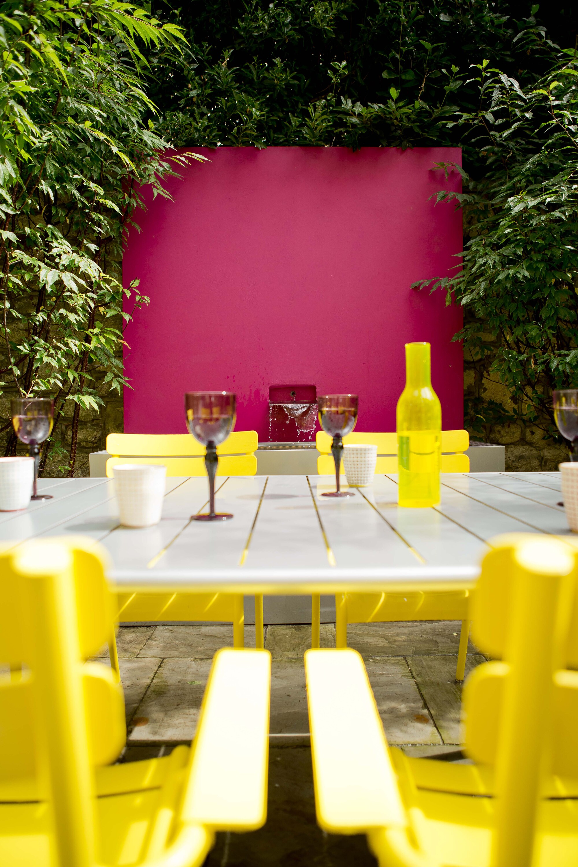An outdoor space with a dining table with bright yellow chairs and a pink back panel as a focal point