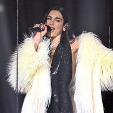 Dua Lipa performs at the 2026 BRIT Awards wearing a sparkly black plunging body suit, a white fur coat, and knee-high boots with fishnets