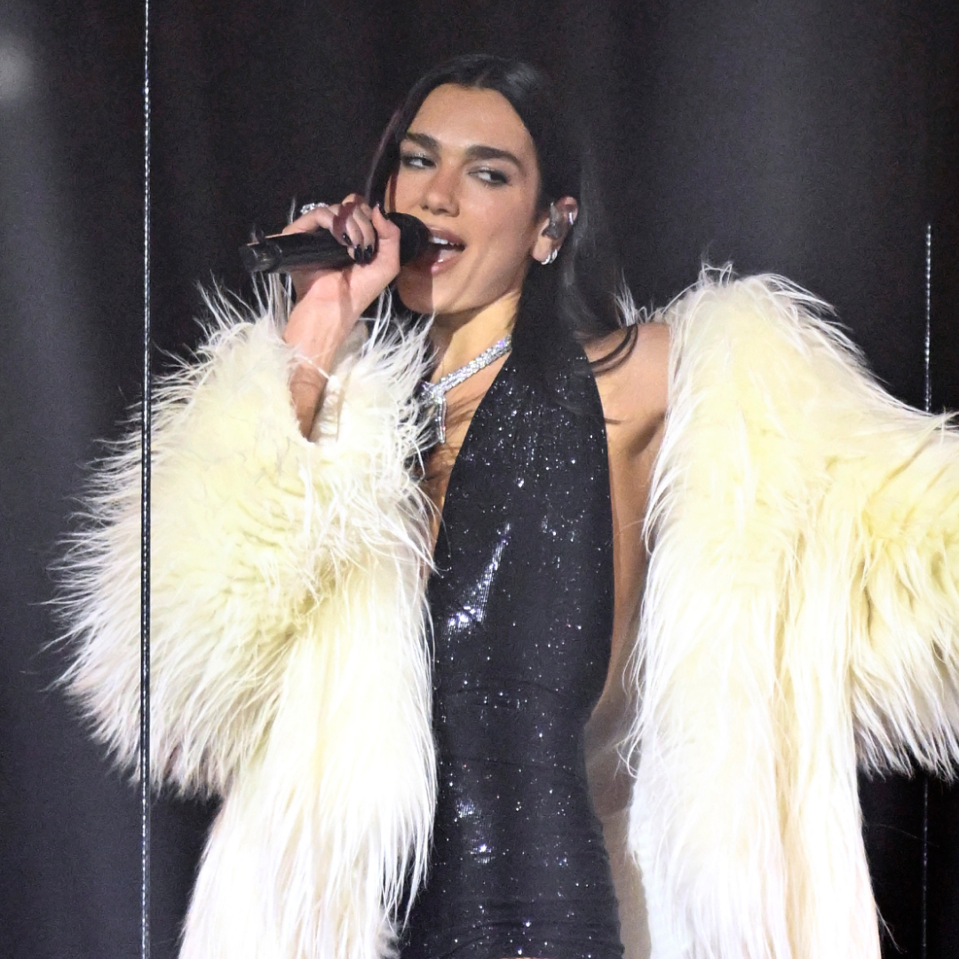 Dua Lipa performs at the 2026 BRIT Awards wearing a sparkly black plunging body suit, a white fur coat, and knee-high boots with fishnets