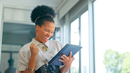 A woman makes a celebratory gesture as she looks at her tablet in an office corridor.