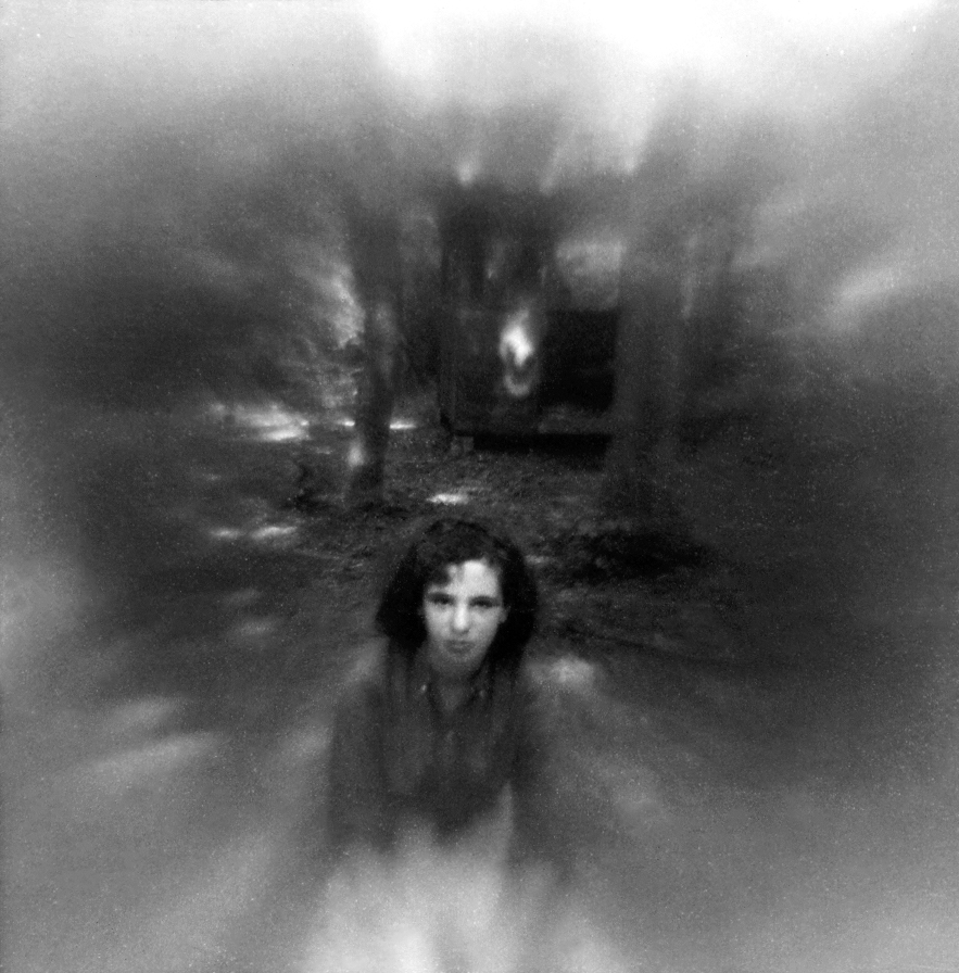 A grainy, atmospheric black and white photograph featuring a person&amp;rsquo;s face centered at the bottom, surrounded by heavy blur and dark trees.