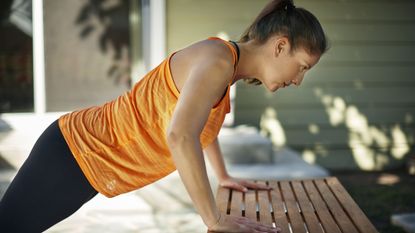 Woman performs incline push-up worth her hands on the seat of a wooden bench