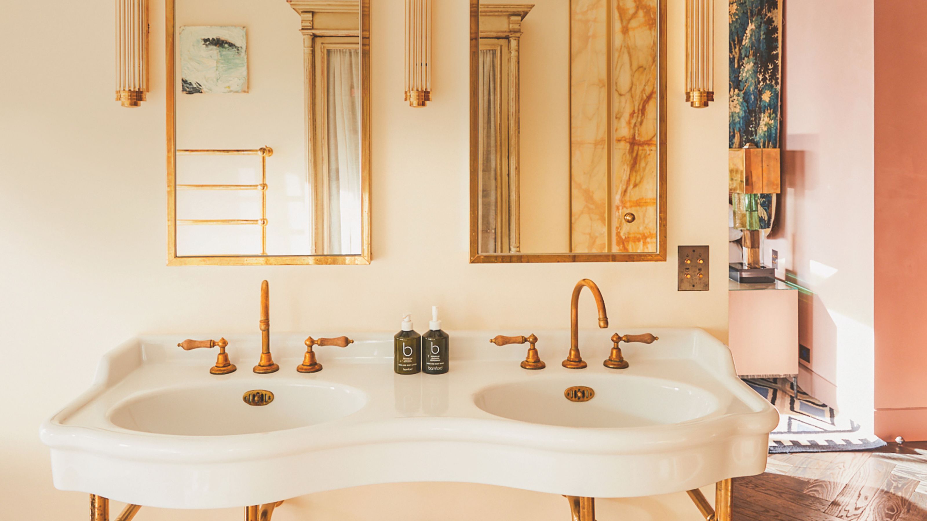 White porcelain double vanity with vintage brass faucets and gold-framed mirrors against cream walls