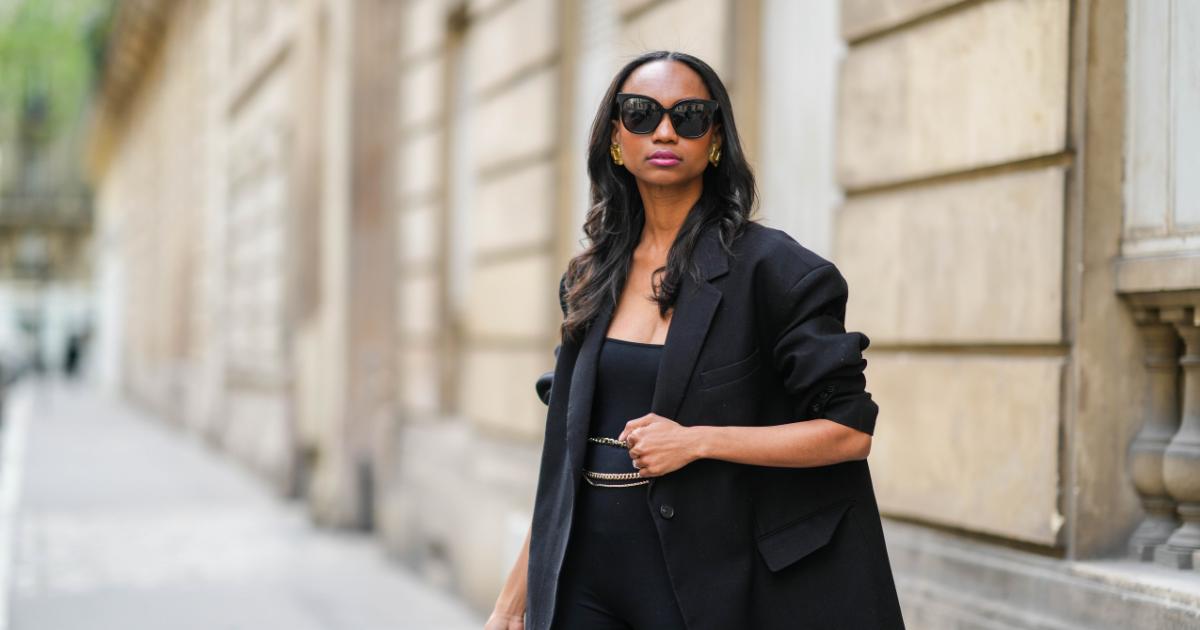 Silk Press Emilie Joseph @in_fashionwetrust wears black sunglasses, gold large vintage earrings, a black oversized blazer jacket from Frankieshop, a black shoulder-off cyclist short jumpsuit, a gold chain belt, gold rings, transparent hair elastics, during a street style fashion photo session, on April 18, 2022 in Paris, France. 