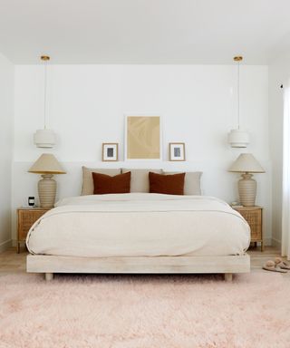A white bedroom with a cream bed, two pendant lights and two tale lamps on wooden nightstands either side, and a peach pink fluffy rug in front