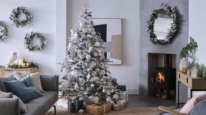 A Scandinavian-style grey and white living room with snowy faux Christmas tree, trio of wreaths on wall and traditional fireplace with flume