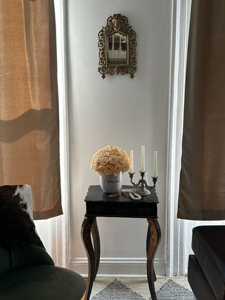 a wooden side table decorated with a candelabra and roses with a vintage mirror on the wall behind and closed beige curtains over the windows either side