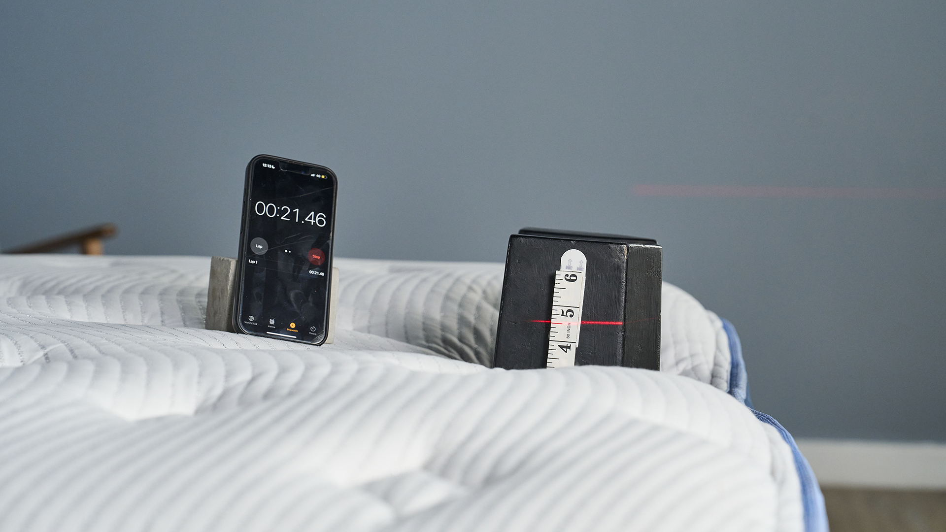 A close-up shot showing the setup for a standardized mattress compression test. A black, rectangular weight with a measuring tape attached is resting on the edge of the white Bear Elite mattress surface. A red laser line is projected across the weight, and a nearby smartphone displays a stopwatch reading of 00:21.46. This setup measures the amount of edge sinkage.