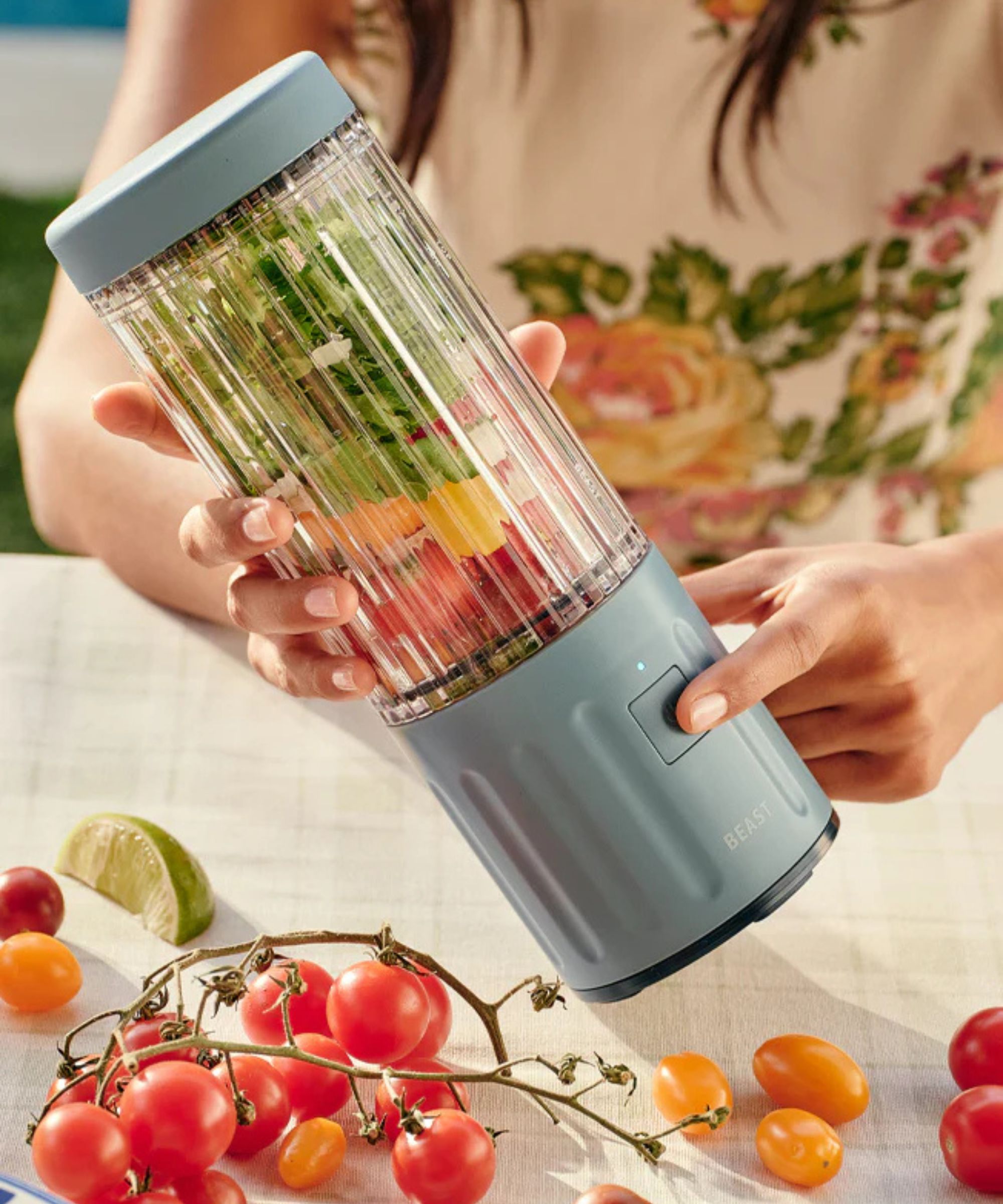 A Caucasian lady holding a small grey portable blender with a clear vessel with salsa ingredients inside, over a picnic table.