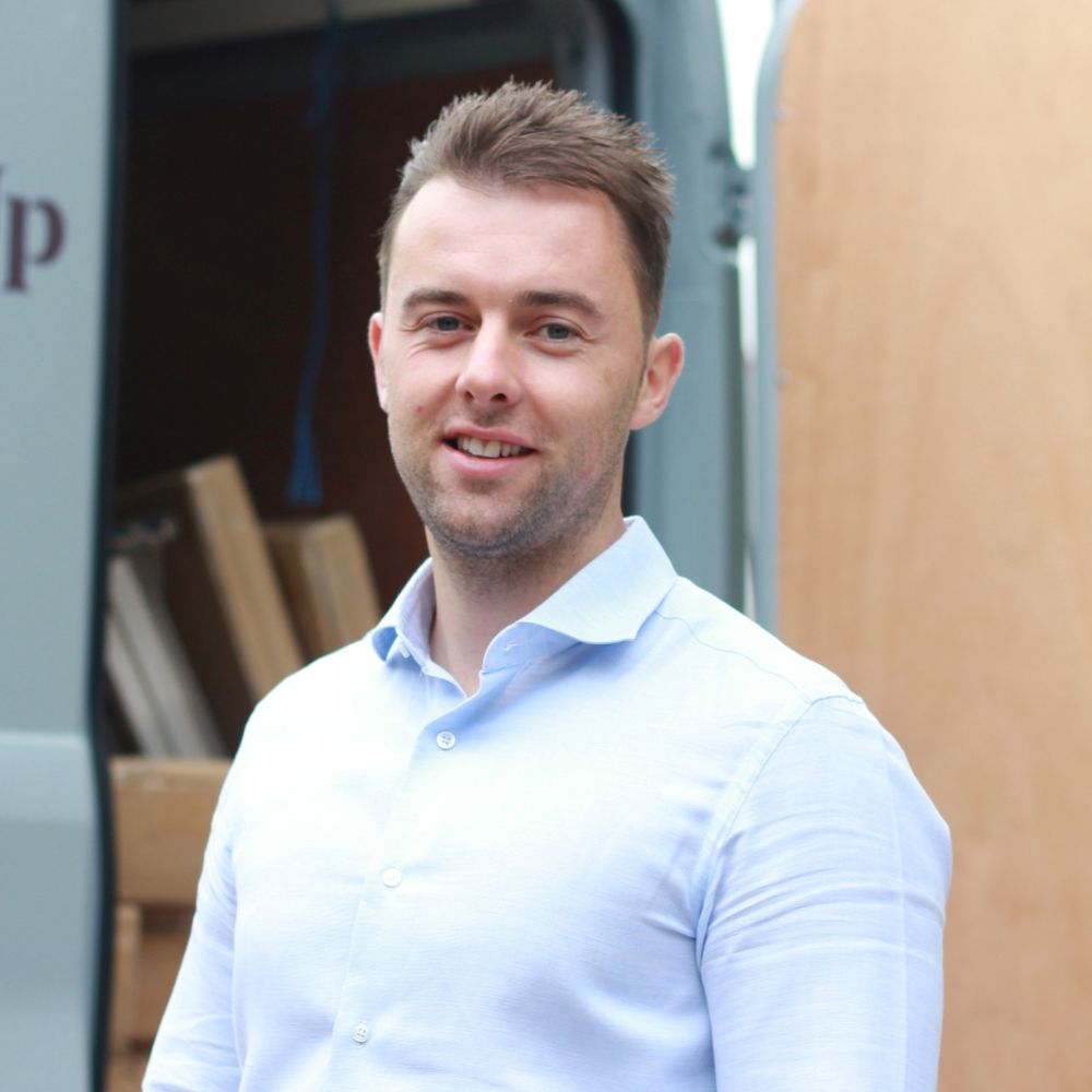 A smiling young man with short brown hair and light stubble, wearing a pale blue shirt, standing in front of an open van loaded with timber boards.