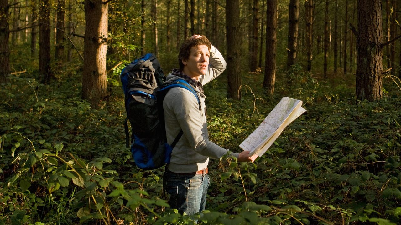 A man hiking in the woods holds a map and looks around, clearly lost.