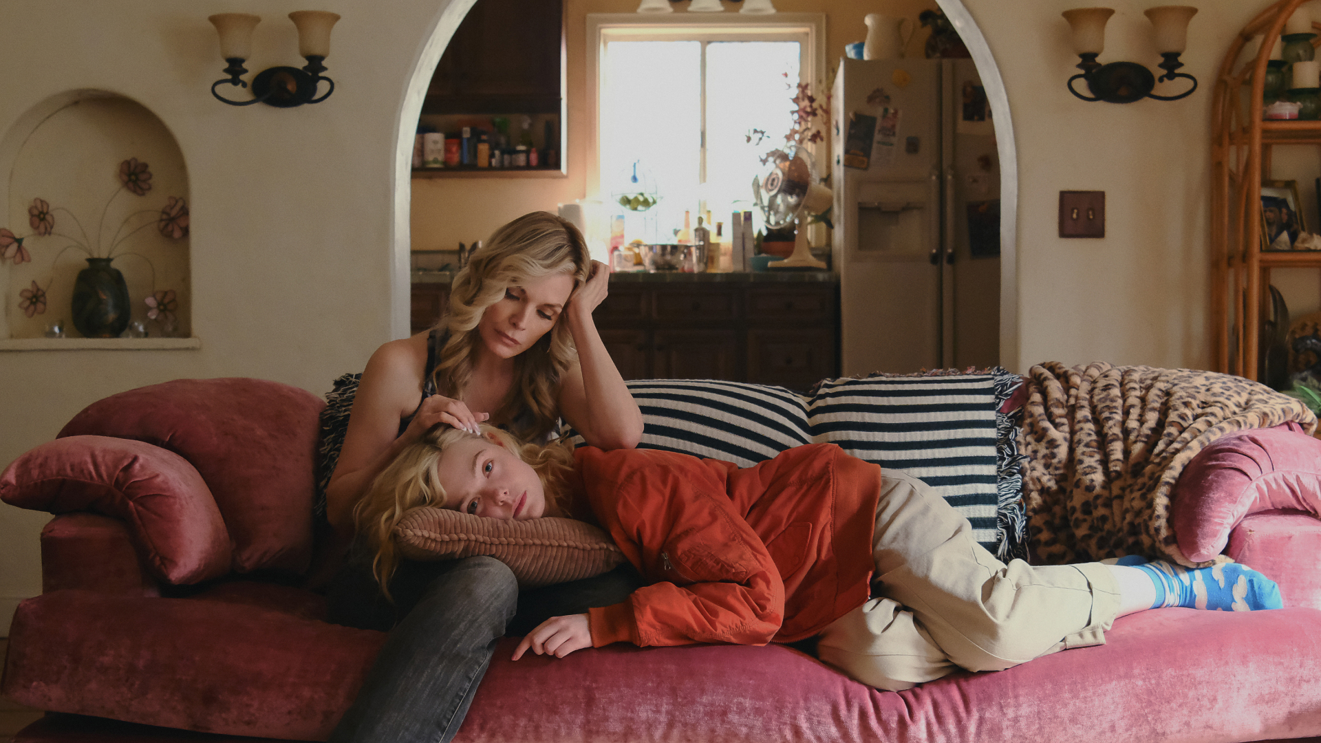 A blonde woman (Elle Fanning) lies on a pink couch with black-and-white striped pillows, resting her head on a pillow in her mother's (Michelle Pfeiffer) lap. The pair sit in a living room with a kitchen behind them, through a circular doorframe, in Apple TV+'s 'Margo's Got Money Troubles.'