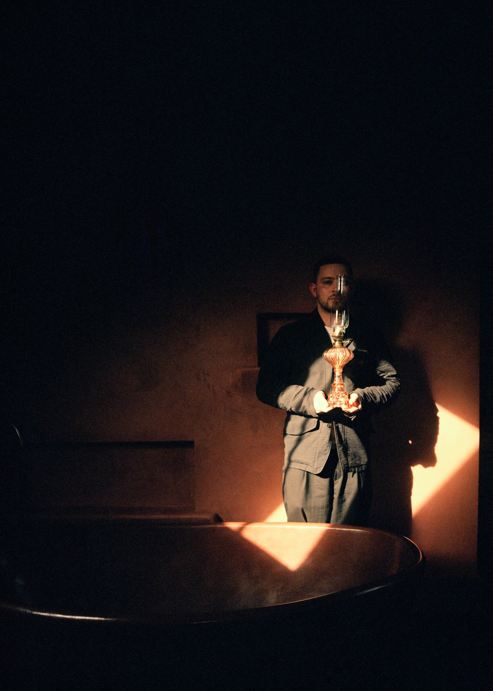 Artist Jobe Burns stands in the corner of a dark room with light reflecting onto him as he holds a glass candle holder up to his face for artistic value